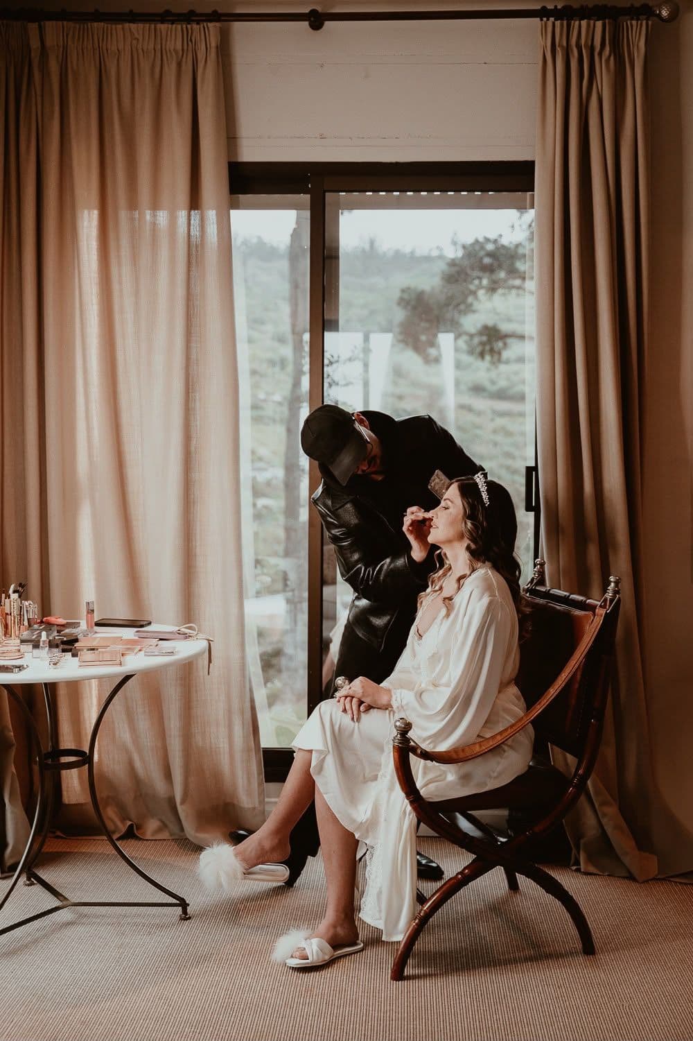 Andony maquillando a una novia junto a la ventana la mañana de su boda